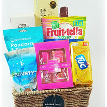 A gift basket hamper containing various sweet foods and grooming products, including chocolates, cookies, a sparkling drink, popcorn, a body spray gift set, a fragrance diffuser, and a granola bar.