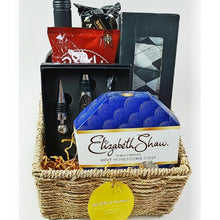 A gift basket containing a variety of items including a bottle of red wine, a box of chocolates, socks, cashews, a wine opener set, and other assorted treats, all arranged in a wicker basket with decorative tulle and a ribbon.