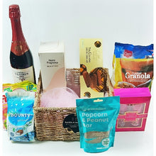 a bottle of St laure red drink, bounty chocolates, Fruitella sweets,Popcorn & Peanut snack, A gift set, Shortbread and Granola all displayed by a handwoven basket all on a white background