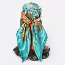 Square silk scarf featuring a vibrant teal and emerald leaf tree design on a golden-brown background with a dark teal border, laid flat on a surface.