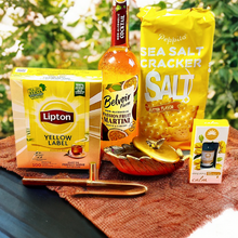 Assorted items including Lipton Yellow Label tea, Belvoir Martini, and Peppino Sea Salt Crackers on a textured surface with greenery in the background.