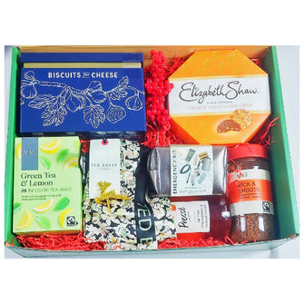 A gift hamper containing a variety of packaged food items and a silk scarf, all neatly arranged in a green box with a red ribbon.