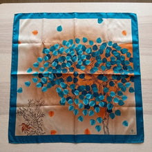 Square silk scarf featuring a vibrant teal and emerald leaf tree design on a golden-brown background with a dark teal border, laid flat on a surface.
