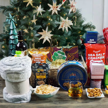 Christmas tree behind a Christmas gift basket set up showing baskets contents. A blanket, cookies, scented candles, crackers, fruit juice, whiskey and red wine paired with gourmet treats like cashews, coconut chips and Hallandais fabric 
