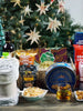 Christmas tree behind a Christmas gift basket set up showing baskets contents. A blanket, cookies, scented candles, crackers, fruit juice, whiskey and red wine paired with gourmet treats like cashews, coconut chips and Hallandais fabric 
