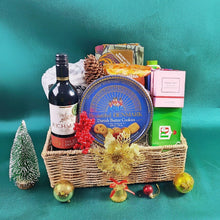 Medium hand woven basket set on a green background with items in the basket such as fabric, a cozy blanket, red wine , whiskey candle, tea, coconut chips and cashews 