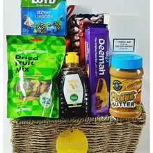 A wicker gift basket filled with assorted snacks and treats, including a green bag of dried fruit mix, a bottle of honey, a purple chocolate bar, a jar of peanut butter, and a blue box of lozenges, with a yellow hanging tag reading 'Ral Gifts & Hampers'.