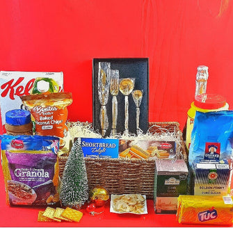 A Christmas hamper containing an assortment of food items including cereal, fruit juice, custard, non-alcoholic drinks, oats, tea, biscuits, chips, and cookies, presented with a golden cutlery set and packaged in a wicker basket against a red background.