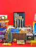 A Christmas hamper containing an assortment of food items including cereal, fruit juice, custard, non-alcoholic drinks, oats, tea, biscuits, chips, and cookies, presented with a golden cutlery set and packaged in a wicker basket against a red background.