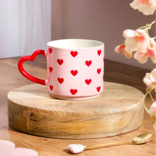 Pink mug with red heart patterns on a wooden stand, surrounded by flowers and a spoon.