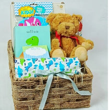 A medium blue themed handcrafted basket  with baby gifts placed in. A teddy, flannel, bath mitts, A T-rex night light and a baby photo frame. 