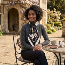 A smiling Black woman with dark curly hair sitting outdoors at a stone bistro table, wearing a dark gray turtleneck sweater and "The Noir & Bloom Silk Scarf" tied neatly around her neck. The black silk scarf features a dramatic white magnolia floral print with dark green leaves and a white-and-black striped border.