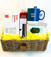A wicker gift basket filled with yellow shredded paper and containing boxed home items, including a blue coffee mug labeled 'COFFEE LOVER,' a glass bottle, an electric wine opener, and a 2-in-1 vacuum cleaner box.