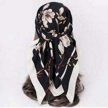 A black square scarf with a white and beige magnolia floral print and brown branch details, styled on a mannequin head with brown hair against a plain white background.