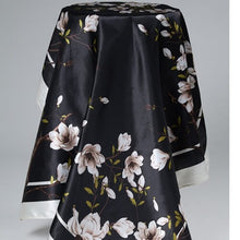 The Noir & Bloom black silk square scarf with white magnolia print, worn by a model in an outdoor lifestyle setting.