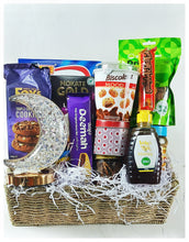 A gift basket containing a variety of snacks including cookies, cornflakes, date bars, latte, fruit juice, sparkling grape drink, incense charcoal, a rechargeable lamp, granola, honey, wax print fabric, mixed dried fruits, and chocolates.