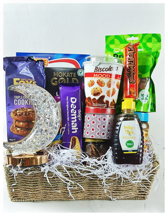 A gift basket containing a variety of snacks including cookies, cornflakes, date bars, latte, fruit juice, sparkling grape drink, incense charcoal, a rechargeable lamp, granola, honey, wax print fabric, mixed dried fruits, and chocolates.