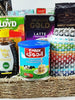 A gift basket containing various food items including tea boxes, biscuits, nuts, honey, and a tea towel, presented in a raffia basket.
