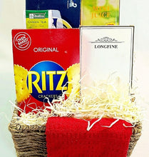 A gift basket arrangement featuring a red Ritz crackers box, a blue and green tea box with a portable juicer, a white Longfine diffuser box, shredded paper filler, and a folded red tea towel in a woven basket.