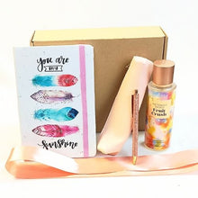 A gift set featuring a white notebook with colorful watercolor feathers and the words 'you are by my sunshine', a pink satin ribbon, a pink body splash bottle labeled 'Victoria's Secret Body Butter Fruit Crash', a pink pen, and a brown gift box.