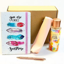 A gift set featuring a 'you are my sunshine' feather notebook, a Victoria's Secret Fruit Crush body spray, a pink purse, and a glittery rose gold pen, arranged in front of a brown cardboard box on a white background.