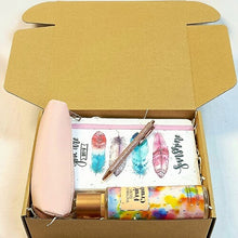 An open kraft cardboard gift box containing a light pink zip pouch, a notebook with colorful feather illustrations and the word 'youre my sunshine', a shimmery metallic pen, and a bottle of body fruit crush splash.
