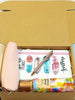 An open kraft cardboard gift box containing a light pink zip pouch, a notebook with colorful feather illustrations and the word 'youre my sunshine', a shimmery metallic pen, and a bottle of body fruit crush splash.