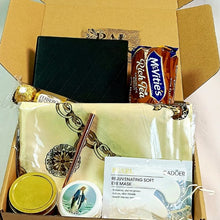 An open cardboard gift box containing multiple items: a black textured wallet, a packet of Rich Tea chocolate biscuits , a gold-wrapped chocolate, a cream-colored scarf with ornate gold chain print, a pen, a round tin candle with a gold lid, a small oval tin with a Mary the Mother of Jesus illustration, and a packaged eye mask labeled Cadoër.