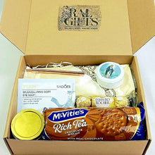 A curated gift box from Ral Gifts containing a McVitie's Rich Tea biscuit, a tin candle, a Sadoer eye mask, a praying rosary in a round case, a pen, scarf , notepad and Ferrero Rocher chocolates, all arranged inside a brown cardboard box.