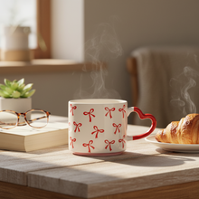 Steaming mug with red bow pattern on a wooden table with a book and croissant in the background.