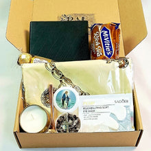 An open cardboard gift box containing a black textured notepad, a ferrero chocolate bar, a cream-colored soft eye mask in plastic packaging, a small white candle, and a round decorative rosary.