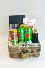 A woven straw gift basket filled with assorted packaged items, including green Heineken cans, a yellow and red Dr Pepper can, a white bag labeled Raba, a black rectangular box, and a black cylindrical container, arranged in white shredded paper inside a natural wicker basket with a hanging gold tag.