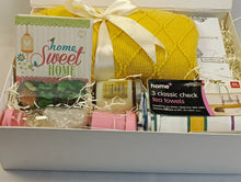 A home gift basket set in a white box, featuring a yellow knitted blanket tied with a white satin ribbon, a 'home Sweet HOME' greeting card, a pack of checkered tea towels, pink portable juicer a small box of pot pourri, glass jar scented candle , and a reed diffuser 