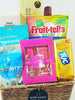 A gift basket hamper containing various sweet foods and grooming products, including chocolates, cookies, a sparkling drink, popcorn, a body spray gift set, a fragrance diffuser, and a granola bar.