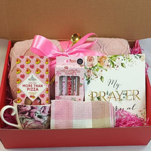 A comfort & prayer inspired gift box set in a red cardboard box, featuring a pink knitted blanket tied with a pink ribbon, a floral mug, a box of Bonbons More Than Pizza chocolates, a ballpoint pen set, and a floral prayer journal 