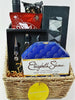 A gift basket containing a variety of items including a bottle of red wine, a box of chocolates, socks, cashews, a wine opener set, and other assorted treats, all arranged in a wicker basket with decorative tulle and a ribbon.