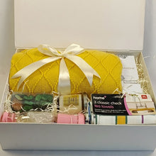 A white gift box filled with assorted household items, prominently featuring a bright yellow knitted throw wrapped in a cream satin ribbon. Other visible items include a pack of tea towels, a boxed product labeled 'home 3 classic check tea towels', a portable juicer, a jar of scented candle , a fragrance diffuser and an apple potpurri