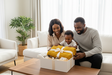 A family of three sits on a white sofa in a bright living room, smiling around a white gift box containing a mustard yellow knitted blanket and gift items.