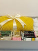 A white gift box filled with assorted household items, prominently featuring a bright yellow knitted throw wrapped in a cream satin ribbon. Other visible items include a pack of tea towels, a boxed product labeled 'home 3 classic check tea towels', a portable juicer, a jar of scented candle , a fragrance diffuser and an apple potpurri