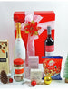 A luxury gift box containing a bottle of Veuve de Verney and Einsberg Non Alcoholic Cabernet Sauvignon, Hareem Istanbul Reed Diffuser, Cartright & Butler Rolled Oat Crumble Biscuits, Cartright & Butler Breakfast Tea, Bayara Mixed Nuts, pine cones, and berry picks, all presented against a red gift box with a decorative tree and gifts.