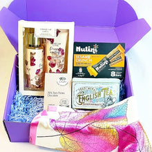 A curated gift box set in a purple box, featuring a '' body splash and hand cream set, a box of Nutini Sesame Crunch bars,  loom chocolate bar, a box of Traditional English Tea, and a pink and yellow patterned silk scarf.