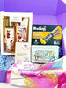 A curated gift box set in a purple box, featuring a '' body splash and hand cream set, a box of Nutini Sesame Crunch bars,  loom chocolate bar, a box of Traditional English Tea, and a pink and yellow patterned silk scarf.