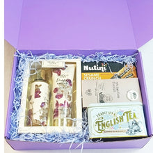 A curated gift box set shown from above inside an open lavender box filled with blue shredded paper. The set includes a floral-patterned bath and body set, a box of Nutini Sesame Crunch cookies, a box of English Breakfast tea, and a tin of Traditional English Tea.
